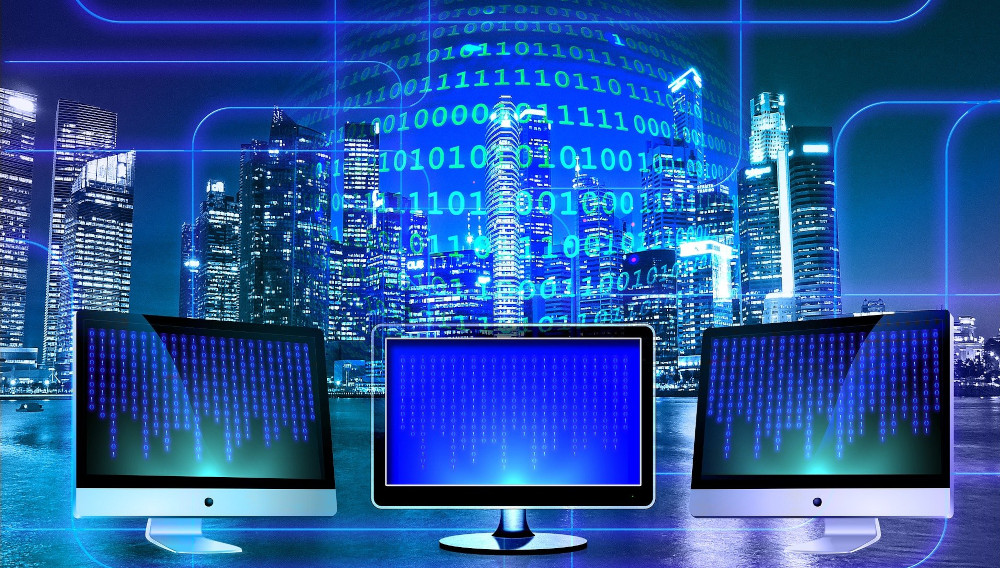 Three computer screens on a desk in front of a window showing a city skyline at night and electric blue numbers and lines (Picture: Geralt on Pixabay)