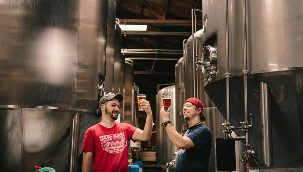 Two people standing in front of fermentation tanks and looking at beer in glasses with a scrutinizing gaze (Photo: Elevate on Unsplash)