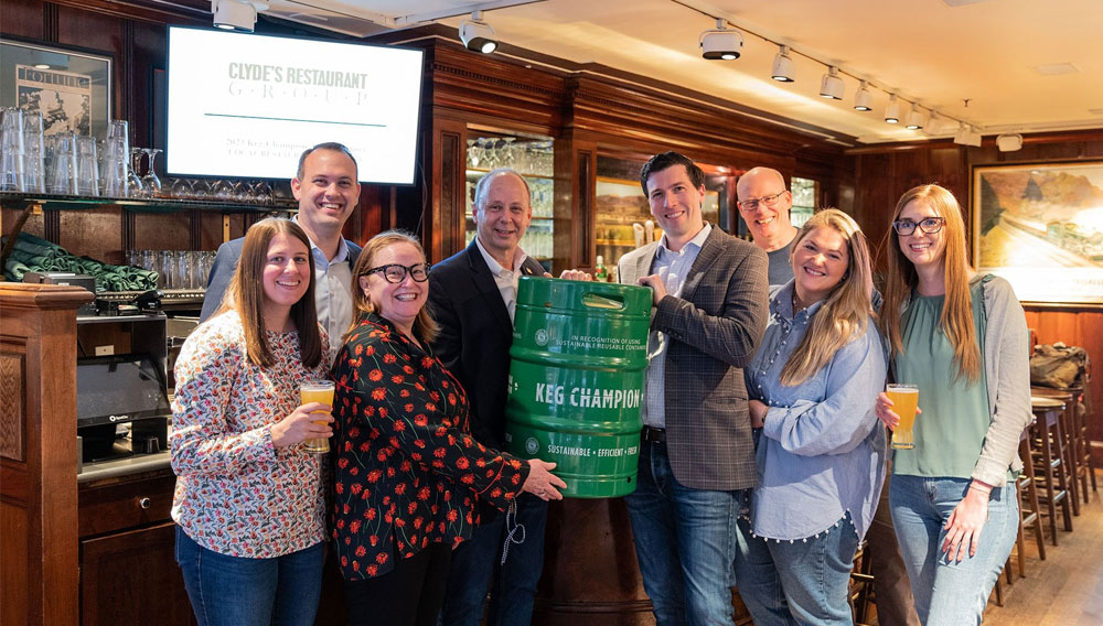 Eight people at the bar of a restaurant, surrounding a green Keg with the words Keg Champion on it, smiling and looking at the camera. In the background, there’s a screen saying, Clyde’s Restaurant