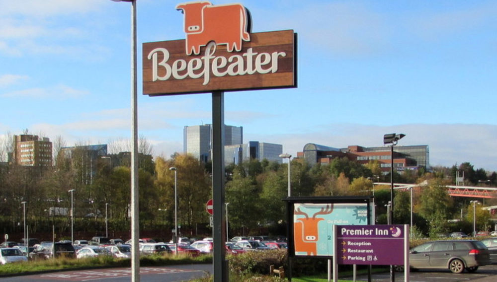 View of a sunny parking lot with a beefeater sign and a beefeater cow on an advertising board in the background (Photo: Beefeater name sign, Euston Way, Telford by Jaggery, https://commons.wikimedia.org/wiki/File:Beefeater_name_sign,_Euston_Way,_Telford_-_geograph.org.uk_-_4780630.jpg, „Beefeater name sign, Euston Way, Telford - geograph.org.uk - 4780630“, https://creativecommons.org/licenses/by-sa/2.0/legalcode)