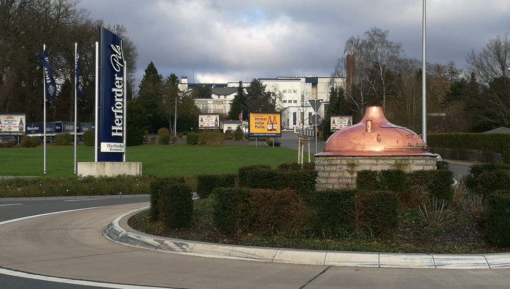 View of a roundabout with a copper kettle in the centre; Herforder flags are flying on the left of the picture; in the background, you can see the Herforder Brewery’s administrative and production buildings (Photo: P6G47TG, https://commons.wikimedia.org/wiki/File:Herforder_Brauerei.jpg, „Herforder Brauerei“, https://creativecommons.org/licenses/by-sa/4.0/legalcode)