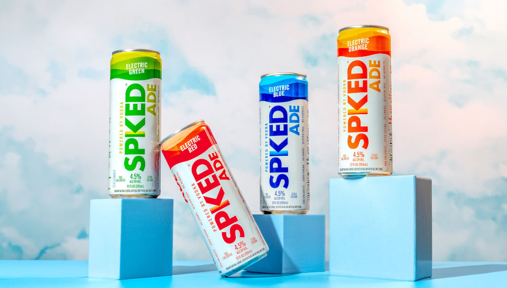 One green, one red, one blue and one orange SPIKED ADE can each placed on light blue surfaces of varying heights, against a background of a light blue sky with white clouds (Photo courtesy of Constellation Brands)