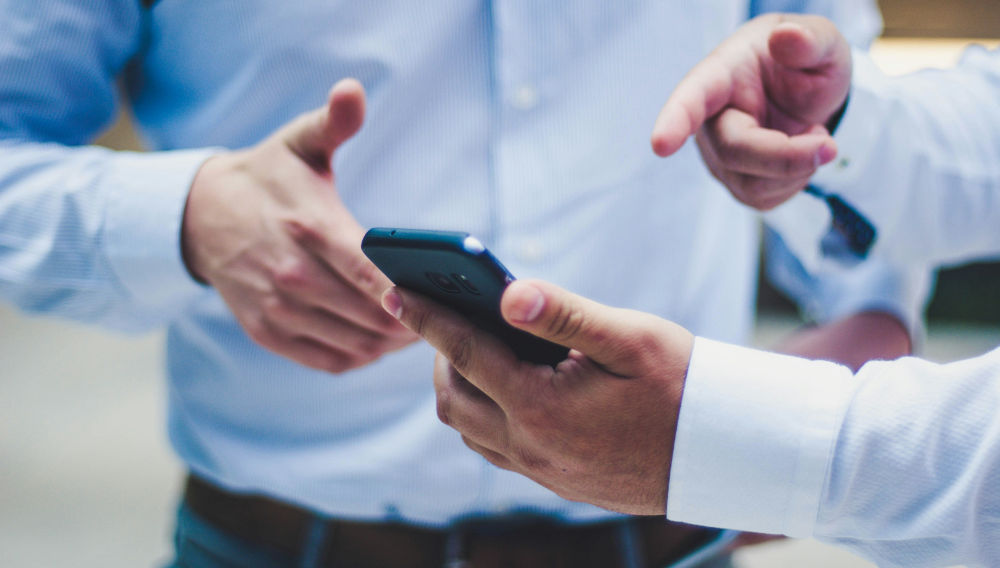 Two people in light blue shirts; it is a close-up, and only their torsos and hands are visible; the person on the right is pointing with one hand at a black mobile phone which they are holding in their other hand (Photo: Luis Villasmil on Unsplash)