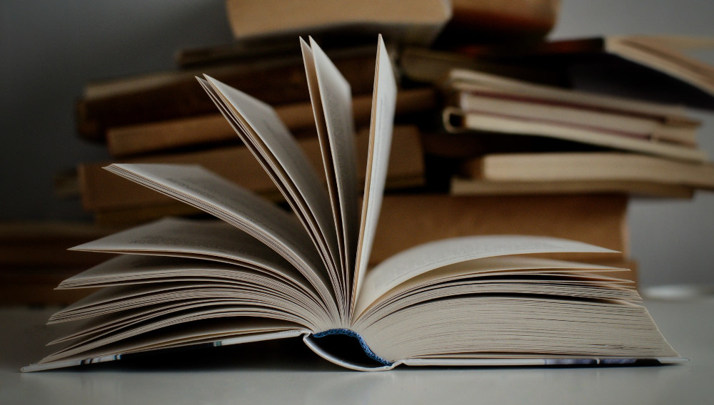 A book lies open on a table, with more books piled up in the background (Photo: Mikolaj on Unsplash)