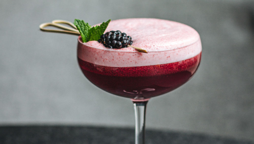 Cocktail glass with raspberry-coloured liquid, garnished with a raspberry and mint leaf (Photo by Nikita Tikhomirov on Unsplash)