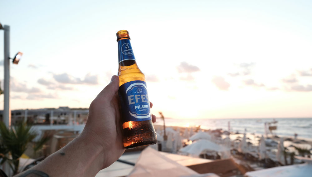 Person hält eine Flasche Efes vor einem sonnenbeschienenen Himmel (Foto: Can Yilmaz auf Unsplash)