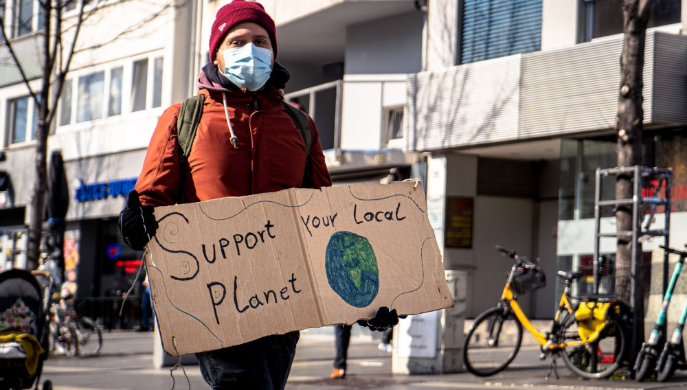 Man carrying a “support your local planet” sign (Photo by Mika Baumeister on Unsplash)