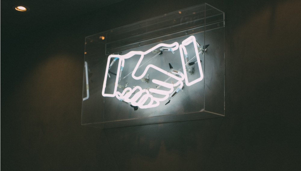 neon sign of a handshake (Photo: Charles Deluvio, Unsplash)