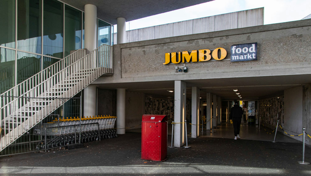 Entrance to a Jumbo Food Market in the Netherlands; On the left-hand side of the picture you can see shopping carts neatly lined up under a staircase leading upwards; in the middle of the picture the supermarket opens up below street level. The building is made of gray concrete, directly in front of the entrance is a red trash can in the middle, above the entrance is the logo of the Jumbo market in yellow. You can see people from behind as they walk further into the market proper (Photo: Denise Jans on Unsplash)