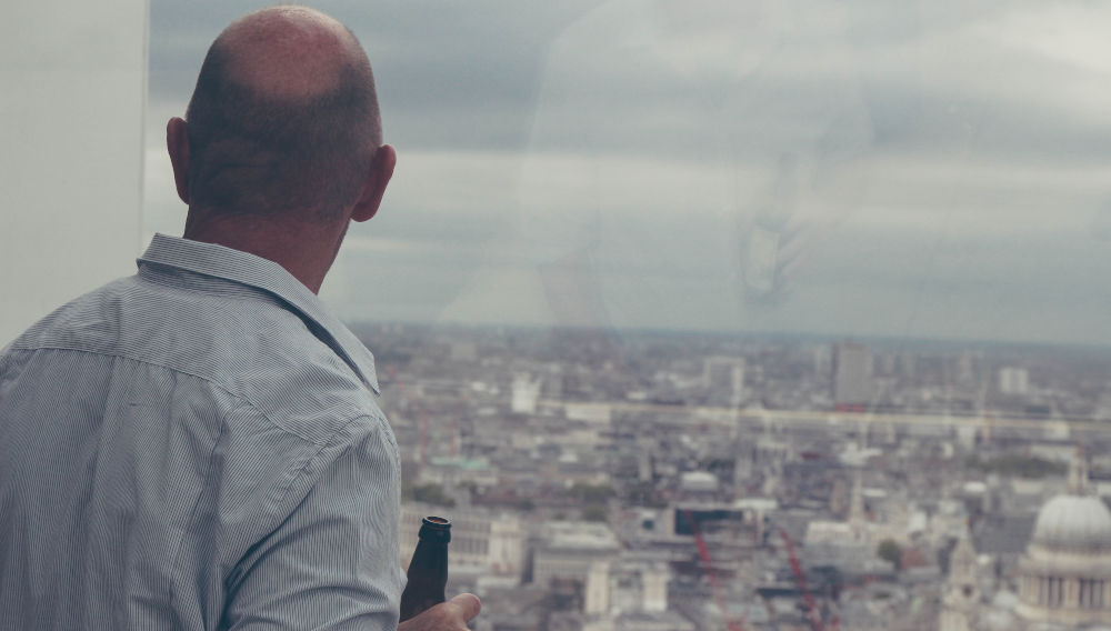 Person in einem Businesshemd hält eine grüne Flasche in der Hand und bewundert das Londoner Panorama durch ein Fenster im Obergeschoss (Foto von Michał Parzuchowski auf Unsplash)