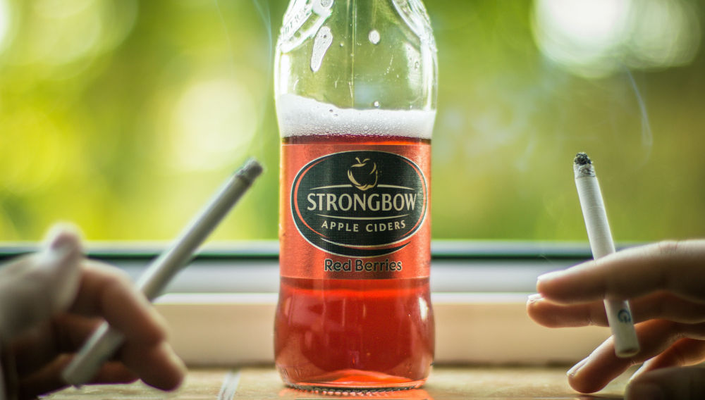 Half empty Strongbow bottle flanked by two hands holding cigarettes (Photo by Haseeb Jamil on Unsplash)
