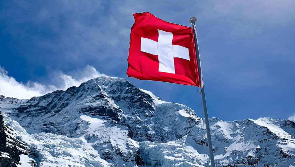 Rote Fahne mit weißem Kreuz auf einem schneebedeckten Berg (Foto: Ronnie Schmutz auf Unsplash)