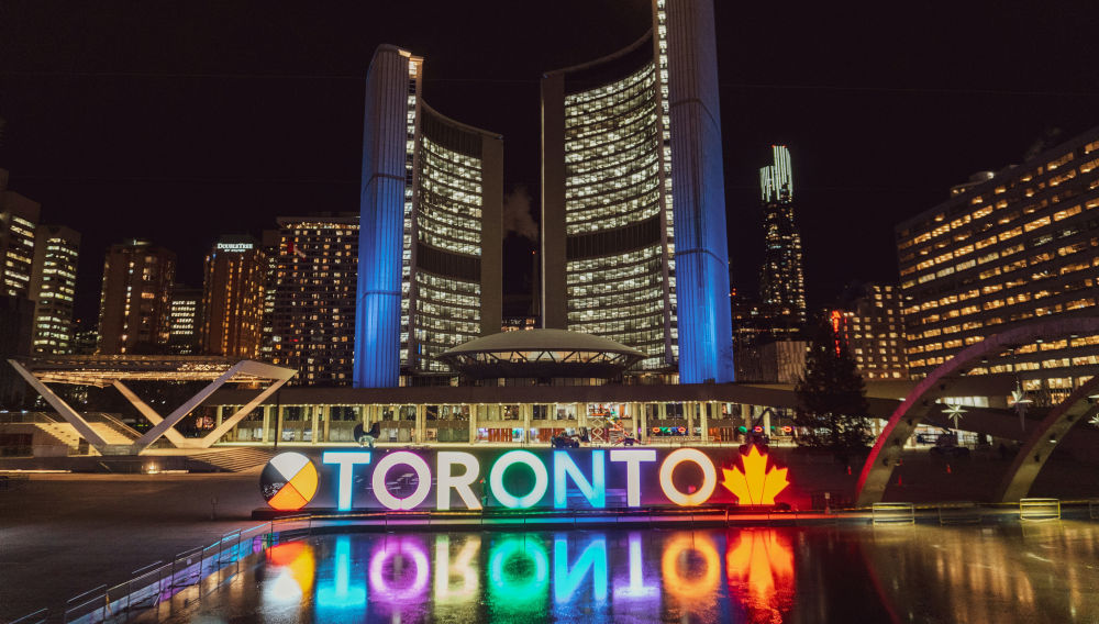 City panorama at night; brightly lit modern skyscrapers against a black night sky in the background; in the foreground we see a Toronto sign in colourful neon letters, reflected in the water (Photo: Conor Samuel on Unsplash)