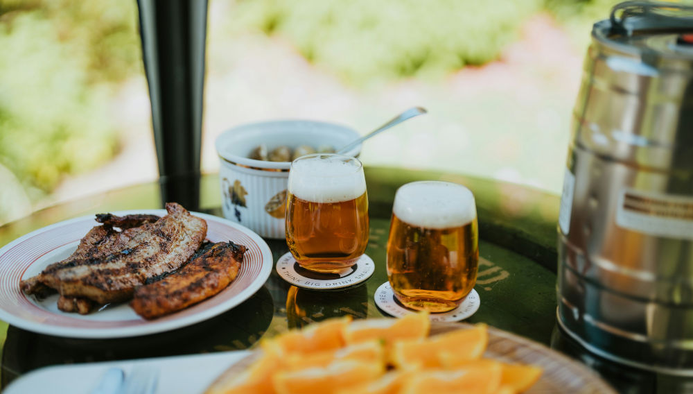 Two half-full beer glasses on a table, next to them are plates of food, on the left a plate of fried meat, on the right edge of the picture you can see an aluminium drinks keg. (Photo: Small Beer on Unsplash)