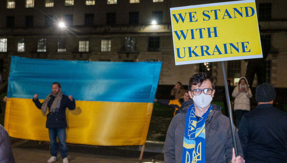 Person holding a blue and yellow protest sign saying “Stand with Ukraine” (Photo: Akhere Unuabona on Unsplash)