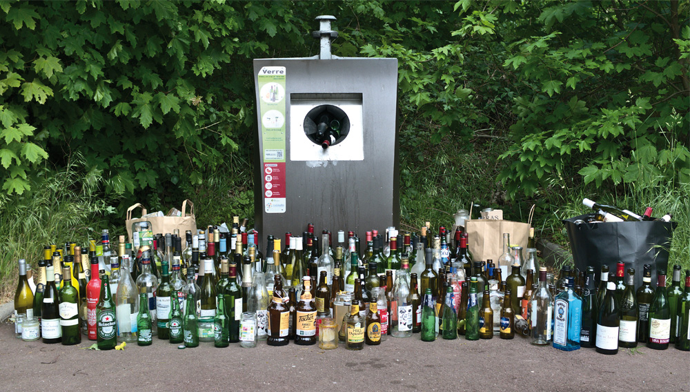 Einweg-Glasflaschen stapeln sich vor einem Recyclingcontainer (Foto: Aleksandr-Kadykov auf Unsplash)