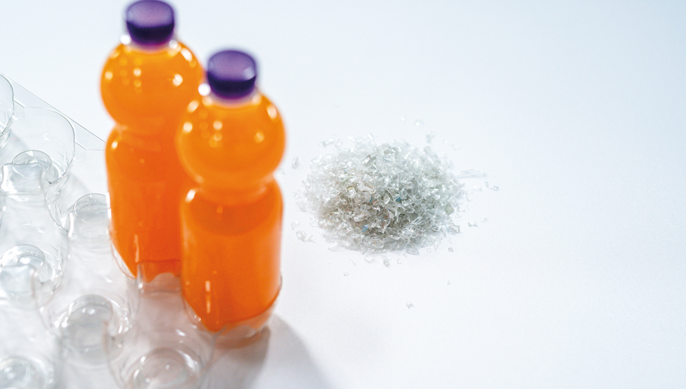 PET-Flaschen mit oranger Flüssigkeit gefüllt, daneben ein kleiner Haufen rPet Recyclingmaterial (Foto: Greiner Packaging)