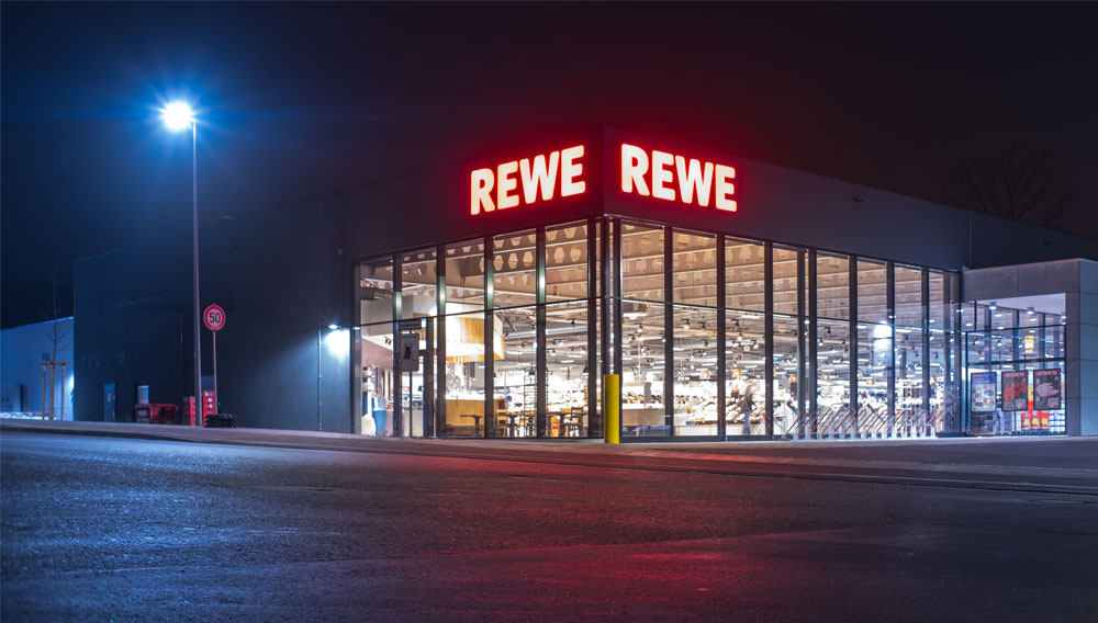 Beleuchteter Rewe-Markt bei Nacht (Foto: Michael Fortsch auf Unsplash)