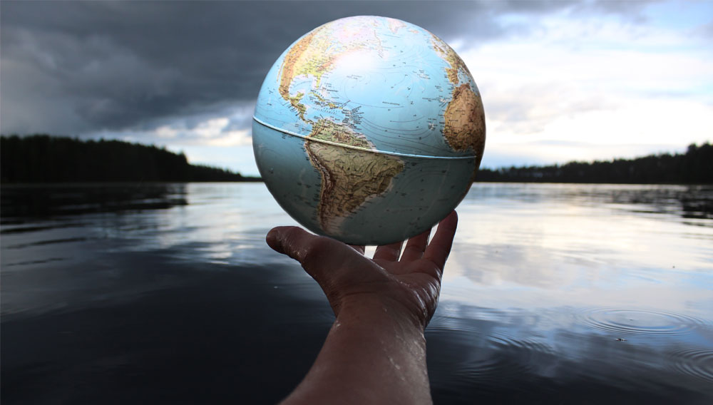 Eine Hand, die einen Globus hält vor dem Hintergrund eines Sees (Foto: Anne Nygard on Unsplash)