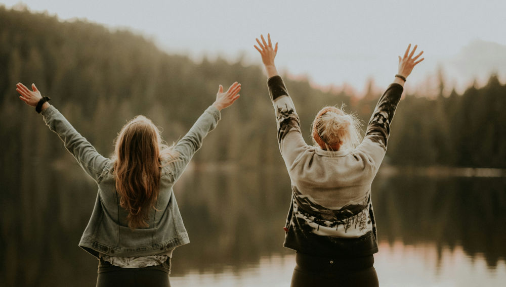 Zwei Menschen stehen mit erhobenen Händen vor einem See (Foto: Priscilla Du Preez auf Unsplash)