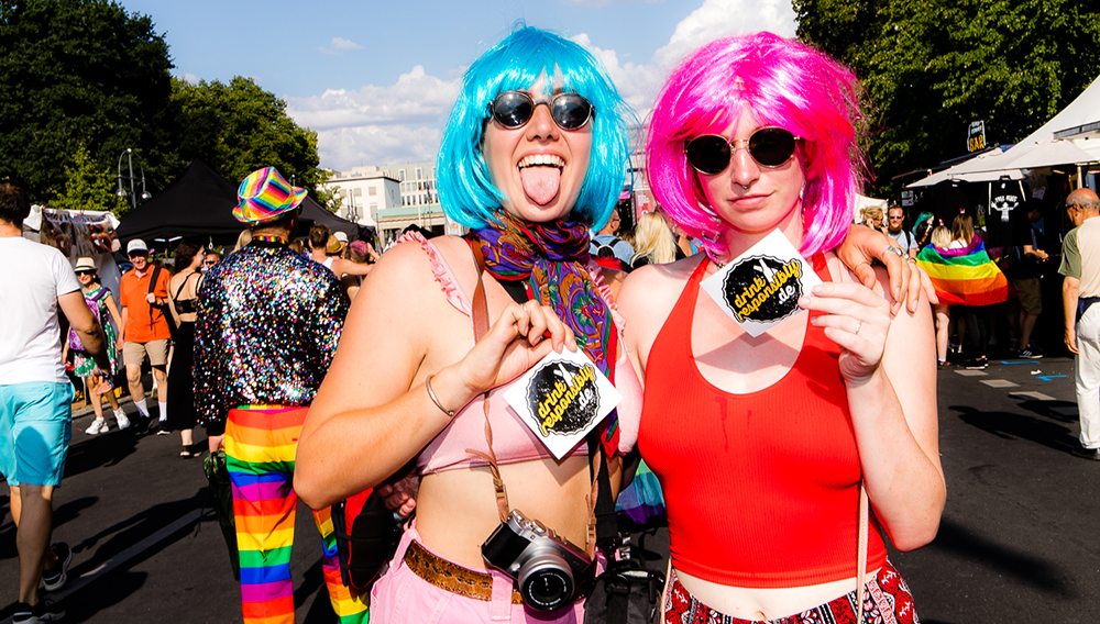 Zwei Frauen mit bunter Kleidung und Perücken halten Flyer mit der Aufschrift Drink responsibly in die Kamera