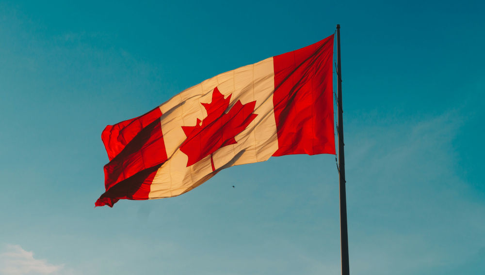 Eine kanadische Flagge weht im Wind, dahinter ist ein hellblauer Himmel zu sehen (Foto: Hermes Rivera auf Unsplash)