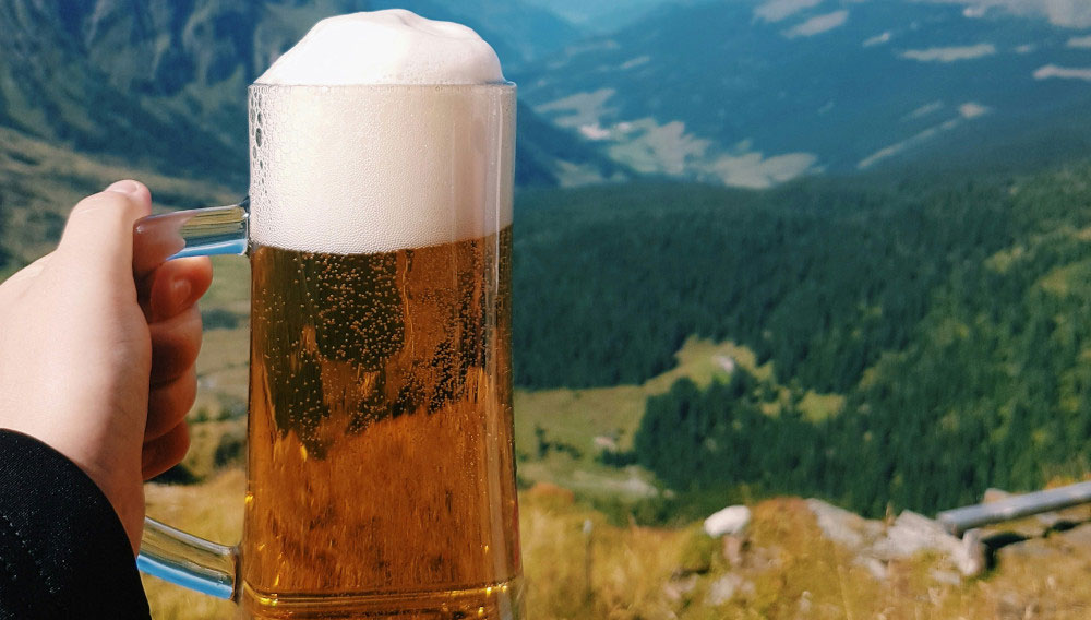 Hand mit Bierglas vor Bergpanorama (Foto: Reiseuhu auf Unsplash)
