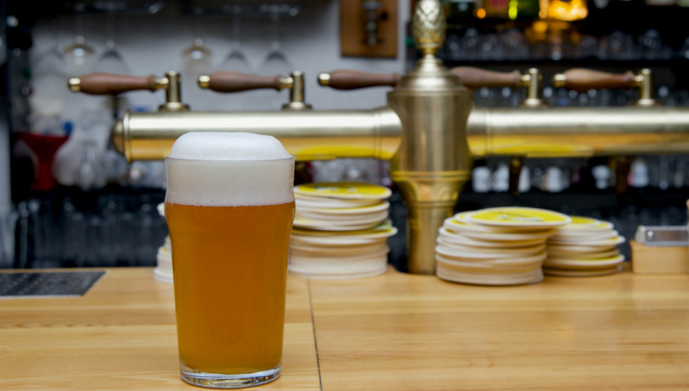 Ein Glas mit trübem Bier und Schaumkrone auf einer Theke vor einer Messing-Schankanlage in einem Gastronomiebetrieb